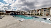This two-floor residential community is centered around a generously scaled outdoor amenity area designed for leisure and social interaction. The centerpiece is a large, resort-style swimming pool with crystal-clear blue water, multiple entry points via stainless steel handrails, and built-in in-water lounge chairs that allow residents to relax directly in the pool. The surrounding concrete pool deck provides ample room for sunbathing and is lined with umbrella-shaded seating areas for additional comfort.

Adjacent to the pool, a covered outdoor pavilion serves as a social hub. Constructed with heavy timber columns and a warm wood-paneled ceiling with recessed lighting, the pavilion features a built-in bar-height counter with wall-mounted brackets, ideal for outdoor dining and entertaining. A mounted flat-screen TV adds a recreational element to the space.

Beyond the pavilion, an expansive open-air patio extends the usable outdoor area, furnished with high-top tables and chairs. A pergola-shaded seating zone provides a quieter retreat surrounded by manicured landscaping. The clubhouse building features large floor-to-ceiling glass doors, prominent columns, and a clean white board-and-batten exterior, creating an inviting and modern architectural focal point for the community.
