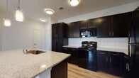 This single-floor residence offers a thoughtfully designed layout with a cohesive modern aesthetic throughout. The kitchen serves as a focal point, showcasing rich dark espresso cabinetry that extends floor-to-ceiling for ample storage, complemented by light granite countertops and a classic white subway tile backsplash. A central island with an integrated sink and pendant lighting above provides additional prep space and a natural gathering point. Black appliances, including an electric range and built-in microwave, complete the kitchen's sleek, contrasting palette. Light wood-style flooring flows through the main living areas, enhancing the sense of openness and warmth. The living area features a ceiling fan, French doors that invite natural light and provide outdoor access, and windows with views of the surrounding landscaping. Additional bedrooms are bright and airy, each with windows that allow generous natural light. The bathroom is finished to a high standard, featuring a wide dual-sink vanity with matching granite countertops and dark cabinetry, large-format tile flooring, a soaking tub, and subway-tiled shower walls. The overall ambiance is clean, contemporary, and move-in ready.