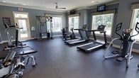 This fitness center offers a comprehensive workout environment designed to support a wide range of exercise routines. The space is finished with durable rubber flooring throughout, providing a safe and resilient surface ideal for both cardio and strength training activities. Large windows line the walls, flooding the room with natural light while offering views of the surrounding outdoor greenery, creating an energizing atmosphere for exercise. The walls are painted in a cool slate blue tone, complemented by classic white crown molding that adds a polished architectural touch. A ceiling fan is centrally mounted overhead, ensuring comfortable airflow during workouts. The fitness center is outfitted with multiple treadmills equipped with individual display screens, a stationary recumbent bike, and a full multi-cable strength training station that supports a variety of resistance exercises. Wall-mounted televisions are positioned for easy viewing from the cardio equipment, enhancing the overall workout experience. Recessed lighting supplements the abundant natural light, keeping the space well-lit at all times. The layout is thoughtfully arranged to allow ample room for movement between equipment, making the fitness center both functional and welcoming.