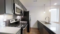 This single-floor space showcases a thoughtfully designed interior with contemporary finishes and a clean, modern aesthetic. The kitchen serves as a standout feature, combining functionality with style. Gray shaker-style cabinetry lines the walls, complemented by a classic gray subway tile backsplash that adds texture and visual interest. Stainless steel appliances, including a smooth-top electric range, over-the-range microwave, and a French door refrigerator, give the kitchen a polished, professional feel. Light-toned countertops provide a crisp contrast against the darker cabinetry, while a stainless steel sink with a modern gooseneck faucet anchors the kitchen island or peninsula. A pendant light fixture hangs overhead, casting focused ambient light over the workspace. Warm hardwood-style flooring flows throughout, adding a natural element that balances the cooler tones of the cabinetry and appliances. Crisp white walls and ceilings enhance the sense of brightness and openness, while ceiling vents suggest a well-integrated HVAC system. The overall ambiance is clean, contemporary, and move-in ready, with every detail lending itself to a comfortable and functional living experience.