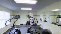 This single-floor fitness center offers a practical and functional workout environment equipped with a variety of cardio machines, including treadmills, ellipticals, and stair climbers. The room is finished with durable, low-pile carpet flooring suited for exercise use, and the walls are painted in a light yellow tone accented by horizontal striped detailing, lending a clean and neutral aesthetic to the space. Overhead fluorescent lighting ensures bright, even illumination throughout the room, complemented by a skylight panel that adds an additional source of light. Large wall-mounted mirrors on one side of the room enhance the sense of space and allow users to monitor their form during workouts. Windows along the walls provide natural light and ventilation, while clearly marked exit doors ensure safety and accessibility. The overall ambiance is straightforward and utilitarian, prioritizing functionality and ease of use for those looking to maintain an active lifestyle.
