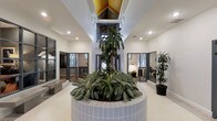 Located at 737 Lockefield Lane in Indianapolis, Indiana, this single-floor space makes an immediate impression with its architecturally distinctive design. At the heart of the layout is a grand atrium-style common area highlighted by dramatically elevated ceilings that open to a skylight with warm wood paneling, flooding the space with natural light. A large, circular tiled planter serves as the centerpiece, housing a mature tropical tree and lush foliage that brings a vibrant, organic element to the interior. The neutral white walls and light flooring create a clean, open backdrop that complements the greenery. Surrounding the central atrium are multiple private rooms enclosed by modern steel-framed glass doors, offering both visibility and separation. These individual rooms are well-lit, with windows allowing natural light to flow in from the exterior. The rooms appear well-suited for office or collaborative use, each furnished with desks and task lighting. Additional potted plants are tastefully placed throughout the space, reinforcing the biophilic design aesthetic. The overall ambiance is professional yet welcoming, blending contemporary architectural details with natural elements to create a functional and visually engaging environment.