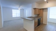 This single-floor space is designed with an open-concept layout that promotes a sense of connectivity and ease of movement. The kitchen is a functional centerpiece, featuring warm wood-toned cabinetry, granite-style countertops, and a peninsula island with an integrated sink — ideal for meal prep and casual dining. A black dishwasher is built into the cabinetry, and the kitchen transitions naturally into the adjacent living area without visual barriers. The living area is finished with plush carpeting that adds warmth underfoot, while a ceiling fan with a light fixture overhead provides both comfort and illumination. Large windows allow natural light to pour into the living space, creating a bright and airy atmosphere throughout. The clean, neutral color palette of white walls and light tones throughout keeps the interior feeling open and versatile, offering a blank canvas easily adaptable to a variety of furnishing styles. The overall layout is practical and well-proportioned, making efficient use of the available space while maintaining a comfortable and welcoming ambiance.