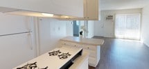 This single-floor residence offers a practical and functional layout centered around an open-concept design that seamlessly connects the kitchen and living areas. The kitchen is outfitted with white cabinetry that extends to the ceiling, maximizing storage capacity, and features a gas cooktop set into a light-toned countertop. A peninsula-style counter provides additional workspace and serves as a natural dividing point between the kitchen and the adjoining living space. The textured ceiling adds a classic residential character throughout the interior. The flooring transitions to warm wood-tone laminate or hardwood in the living area, lending a cohesive and welcoming feel to the space. The living room benefits from natural light streaming through a sliding glass door dressed with vertical blinds, which also provides direct access to the outdoors. A wall-mounted HVAC unit ensures climate control convenience. The overall palette of whites and neutral tones creates a clean, airy ambiance, making the space feel open and easy to personalize. The straightforward single-floor layout offers ease of navigation and accessibility throughout.