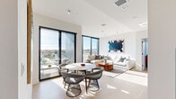This thoughtfully designed one-bedroom, one-bathroom unit showcases a contemporary aesthetic throughout its efficiently laid-out single floor. The open-plan living and dining area is defined by expansive floor-to-ceiling black-framed windows and a sliding glass door that frames sweeping panoramic city views, flooding the space with abundant natural light and light wood-tone flooring underfoot.

The kitchen runs along one wall in a streamlined galley configuration, featuring concrete-finish cabinetry extending to the ceiling, a blue-toned backsplash, stainless steel appliances including a French door refrigerator and gas range, and dark countertops — a striking contrast to the bright, airy living space it adjoins.

The bedroom is a serene retreat with plush carpeting, a large window with urban views, and warm wall-mounted lighting. An adjacent hallway features floor-to-ceiling warm wood-paneled built-in cabinetry with integrated storage, providing practical organization.

The bathroom is finished with speckled terrazzo-style tile flooring, a floating wood-accented vanity, black-framed mirrors with pendant lighting, open glass shelving, and a full soaking tub with a glass-enclosed shower. Clean white walls and recessed lighting contribute to the unit's calm, modern ambiance throughout.