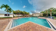 Located at 5100 West Sample Road in Coconut Creek, Florida, this outdoor amenity space showcases a resort-style swimming pool that serves as a central gathering point for the community. The pool features a classic L-shaped design with clear, blue-tinted water and is framed by a generously sized, terracotta-toned brick paver deck that provides ample room for lounging and recreation.

The surrounding environment is richly landscaped with tall royal palms, broad-canopy shade trees, and lush tropical greenery that creates a private, retreat-like atmosphere. Lounge chairs and seating are arranged along the poolside, offering comfortable spaces for relaxation. A secure perimeter fence encloses the area, adding both safety and a sense of definition to the space.

The backdrop includes views of the residential buildings beyond the treeline, blending harmoniously with the natural setting. The open-air design allows for abundant natural light and unobstructed views of the South Florida sky, enhancing the overall outdoor experience. The combination of the vibrant aquamarine pool, warm paver surroundings, and dense tropical foliage creates an inviting, sun-drenched atmosphere characteristic of premier Florida living. This well-maintained outdoor space offers a peaceful and visually appealing escape for residents and visitors alike.