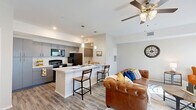 This thoughtfully designed single-floor residence offers a seamless blend of functionality and modern style. The open-concept kitchen and living area serves as the heart of the unit, where wood-look flooring, neutral tones, and abundant natural light from multiple windows create a bright and welcoming atmosphere. A ceiling fan adds to the comfort of the living space, which is anchored by a corner fireplace and media center.

The kitchen is outfitted with sleek grey flat-front cabinetry extending to the ceiling for maximum storage, light stone countertops, a kitchen island with bar seating, pendant lighting, and a full suite of black appliances including a refrigerator, range, and over-the-range microwave. A double sink with additional lower cabinetry lines the opposite wall, keeping the workspace organized and efficient.

The bedroom is a comfortable retreat featuring a ceiling fan, a natural light-welcoming window, and direct access to both a closet and the full bathroom. The bathroom mirrors the kitchen's design aesthetic with grey cabinetry, a single vanity with a wide mirror, and a shower/tub combination. A dedicated laundry area with overhead cabinetry and washer/dryer hookups adds everyday convenience, while a utility room and additional closet provide generous storage throughout.