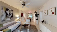 This well-appointed single-floor residence showcases a clean, modern aesthetic defined by an open-concept floor plan that encourages natural movement between living spaces. The living area is anchored by a plush white sofa, a glass-top coffee table, and a textured area rug, all set atop light-toned tile flooring that extends throughout the home. A ceiling fan overhead adds both function and a casual elegance to the space.

The layout flows effortlessly into the dining area, where a dark wood table is paired with white modern chairs beneath a contemporary chandelier, creating a striking contrast. A large mirror in the dining space enhances the sense of depth and reflects ambient light throughout the room.

Adjacent to the dining area, the kitchen features dark cabinetry and a raised breakfast bar with seating, offering a casual alternative for dining. The open pass-through design keeps the kitchen visually connected to the rest of the living spaces.

Thoughtful decorative touches — including a sculptural bonsai tree in a tall white planter, a well-placed table lamp, and curated accessories on a low media console — give the space a cohesive, designed feel. Bright white walls and abundant lighting contribute to an airy, welcoming ambiance throughout.