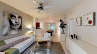 This well-appointed single-floor residence showcases a clean, modern aesthetic defined by an open-concept floor plan that encourages natural movement between living spaces. The living area is anchored by a plush white sofa, a glass-top coffee table, and a textured area rug, all set atop light-toned tile flooring that extends throughout the home. A ceiling fan overhead adds both function and a casual elegance to the space.

The layout flows effortlessly into the dining area, where a dark wood table is paired with white modern chairs beneath a contemporary chandelier, creating a striking contrast. A large mirror in the dining space enhances the sense of depth and reflects ambient light throughout the room.

Adjacent to the dining area, the kitchen features dark cabinetry and a raised breakfast bar with seating, offering a casual alternative for dining. The open pass-through design keeps the kitchen visually connected to the rest of the living spaces.

Thoughtful decorative touches — including a sculptural bonsai tree in a tall white planter, a well-placed table lamp, and curated accessories on a low media console — give the space a cohesive, designed feel. Bright white walls and abundant lighting contribute to an airy, welcoming ambiance throughout.