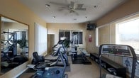This single-level fitness room offers a functional and well-organized workout space designed to accommodate a variety of exercise needs. The room is finished with warm beige walls and textured ceilings, giving it a comfortable, welcoming atmosphere. A ceiling fan is centrally mounted overhead, complementing the recessed lighting to ensure proper ventilation and visibility throughout the space.

Natural light filters in through multiple windows fitted with vertical blinds, brightening the interior and providing a sense of openness. The flooring transitions between carpet and rubber mat sections, with the matted areas strategically placed beneath the strength training equipment for stability and protection.

The room is equipped with a selection of cardio machines, including stationary bikes, ellipticals, and a treadmill positioned along the perimeter. A multi-station cable and weight machine serves as the centerpiece for strength training. A wall-mounted television is installed in the corner, allowing users to stay entertained during workouts. A small shelving unit provides additional storage near the treadmill. The layout is thoughtfully arranged to maximize usable floor space while keeping pathways clear between equipment.