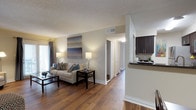 This single-floor residence offers a thoughtfully designed layout that balances comfort and functionality. The open-concept living area serves as the heart of the home, featuring rich hardwood flooring that flows seamlessly throughout the space. Neutral wall tones create a warm, inviting atmosphere, complemented by a textured ceiling and ample natural light streaming in through large windows dressed with elegant curtains.

The living area provides generous space for relaxation and entertaining, with room for a full sofa arrangement and a central coffee table. Recessed lighting and floor lamps contribute to a well-lit, welcoming ambiance. A piece of abstract wall art adds a modern decorative touch to the main living space.

Adjacent to the living area, the kitchen features dark cabinetry with a sleek countertop, visible from a pass-through opening that maintains the open feel of the floor plan. The kitchen is equipped with modern appliances and offers practical counter space for meal preparation.

A hallway extends from the living area, lined with closet doors and leading to the additional rooms of the residence. The consistent hardwood flooring and neutral palette carry throughout, tying the entire space together with a cohesive and polished aesthetic.