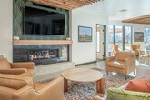 Located at 219 N Fair Oaks Ave in Madison, WI, this contemporary community amenity space spans a generous footprint across its main level, offering a well-appointed interior complemented by an expansive outdoor courtyard. The interior is defined by a striking slatted wood ceiling that adds warmth and architectural character throughout. The main gathering area is divided into distinct zones: a comfortable lounge featuring tan leather sofas and armchairs arranged around a fireplace with decorative tile surround, and a dining area furnished with marble-top tables and wooden chairs beneath globe pendant lights. A built-in bar with an arched wood-clad backdrop, open shelving, and pendant lighting serves as a central focal point. Large floor-to-ceiling glass doors flood the interior with natural light and provide seamless access to the outdoor patio. The outdoor courtyard is equally well-equipped, featuring a built-in stainless steel grill station, a raised bar counter with seating, a fire pit surrounded by colorful Adirondack chairs, and a pergola-shaded gathering area. The multi-story residential building enclosing the courtyard features modern panel cladding, private balconies, and large windows, creating an enclosed yet open-air atmosphere ideal for community gatherings.