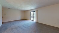 Located at 197 Rosemary St in Needham Heights, MA, this single-floor unit offers a practical and functional layout spread across several interconnected spaces. The living area is generously sized, featuring wall-to-wall blue-gray carpeting, neutral cream-toned walls, and a textured ceiling. A sliding glass door opens to a balcony, allowing natural light to filter in and providing views of surrounding greenery—a notable feature of the main living space. An adjacent area transitions to hard surface flooring and connects to a dining space, which is highlighted by a pendant light fixture overhead.

The galley-style kitchen is outfitted with dark wood cabinetry on both sides, light-toned countertops, and hard surface flooring. It includes a white refrigerator, electric range with oven, built-in dishwasher, and a stainless steel sink, offering efficient use of the space.

The bedroom is carpeted, receives natural light through a window, and features a walk-in closet alongside an additional sliding door closet, providing ample storage.

The full bathroom includes a vanity with a single sink set into a white countertop with decorative cabinetry below, a tub-shower combination with teal surround panels, a small window, and classic mosaic tile flooring in blue and white tones.