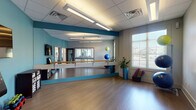 Located at 535 Blackrock Dr in Blacksburg, VA, this single-floor fitness and wellness studio is thoughtfully designed to support a variety of exercise and movement activities. The centerpiece of the space is a spacious group exercise room finished with smooth hardwood-style flooring, vibrant teal accent walls, and a full wall of floor-to-ceiling mirrors. Wall-mounted wooden ballet barres run the length of the mirror wall, making the room well-suited for barre, yoga, Pilates, and similar disciplines. Bright fluorescent lighting panels installed in the drop-tile ceiling ensure the space is evenly and generously lit throughout. Large windows along one wall invite natural light and offer views of the surrounding neighborhood, contributing to an open and energizing atmosphere. Exercise equipment including stability balls, hand weights, and yoga mats are neatly stored within the room. A wall-mounted flat-screen television and a digital kiosk provide instructional and entertainment support during sessions. Interior glass partition windows and frosted-glass doors connect the studio room to adjacent areas, including what appears to be a separate gym or equipment room, promoting a cohesive and interconnected facility layout. The overall design is clean, modern, and functional, creating an inviting environment for fitness and wellness activities.