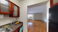 This single-floor unit offers a well-organized layout with a consistent design language throughout. The most striking feature is the rich, honey-toned parquet hardwood flooring that extends seamlessly across the living areas and bedroom, lending warmth and classic character to the space.

The kitchen is compact yet functional, outfitted with rich cherry wood cabinetry featuring frosted glass upper panels and sleek bar-pull hardware. Dark granite-style countertops provide a strong contrast against the cabinetry, and the space includes a stainless steel sink and a gas range, making it a practical cooking environment. A window allows for natural light and ventilation.

The living area is open and generously proportioned, connected to the kitchen via a wide archway that promotes an easy flow between spaces. Clean white walls and natural light from a window create a bright, airy atmosphere. A window-mounted air conditioning unit and a radiator beneath the window provide year-round climate comfort.

The bedroom mirrors the living area's warmth with the same polished parquet floors, a ceiling fan with light fixture, a window for natural light, and a dedicated closet. The room offers a calm and functional retreat.

The bathroom features classic white subway tile wainscoting, a vanity with a brushed nickel faucet, a wall-mounted mirror, and a shower-tub combination — clean and straightforward in design.