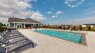 Located on Weymouth Way in Perrysburg, Ohio, this outdoor community pool facility offers a well-appointed recreational space designed for leisure and social gatherings. The centerpiece is a large, rectangular swimming pool with clear blue water, accessible via entry steps and bordered by a broad concrete deck that provides ample space for circulation and relaxation.

The sundeck is lined with numerous chaise lounge chairs arranged along all sides of the pool, offering residents generous options for sunbathing. Shaded dining areas featuring table-and-chair sets with umbrellas are positioned along the perimeter, providing comfortable spots for outdoor meals or casual conversation.

A wood-paneled pergola structure is situated at one end of the deck, adding a distinct architectural element and additional shaded seating. The entire pool area is enclosed by a black metal fence, with manicured evergreen plantings and decorative landscaping enhancing the visual appeal along the boundaries.

Adjacent to the pool, a single-story clubhouse with gray lap siding, white trim, a hip roof, and large windows serves as a complementary facility. Its covered porch faces the pool area, creating a seamless indoor-outdoor connection. The surrounding neighborhood and open sky views contribute to the bright, open ambiance of the space.