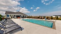 Located on Weymouth Way in Perrysburg, Ohio, this outdoor community pool facility offers a well-appointed recreational space designed for leisure and social gatherings. The centerpiece is a large, rectangular swimming pool with clear blue water, accessible via entry steps and bordered by a broad concrete deck that provides ample space for circulation and relaxation.

The sundeck is lined with numerous chaise lounge chairs arranged along all sides of the pool, offering residents generous options for sunbathing. Shaded dining areas featuring table-and-chair sets with umbrellas are positioned along the perimeter, providing comfortable spots for outdoor meals or casual conversation.

A wood-paneled pergola structure is situated at one end of the deck, adding a distinct architectural element and additional shaded seating. The entire pool area is enclosed by a black metal fence, with manicured evergreen plantings and decorative landscaping enhancing the visual appeal along the boundaries.

Adjacent to the pool, a single-story clubhouse with gray lap siding, white trim, a hip roof, and large windows serves as a complementary facility. Its covered porch faces the pool area, creating a seamless indoor-outdoor connection. The surrounding neighborhood and open sky views contribute to the bright, open ambiance of the space.