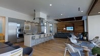 This contemporary single-floor space showcases a thoughtfully designed open-concept layout that effortlessly connects the kitchen and living areas. The kitchen is a standout feature, boasting a generous central island with white bar seating, a stainless steel range hood suspended from the ceiling, and a classic subway tile backsplash that adds texture to the clean, modern aesthetic. Pendant lighting hangs above the island, complementing the recessed ceiling lights throughout. Stainless steel appliances, including a built-in wall oven and refrigerator, complete the well-appointed culinary space. The kitchen flows naturally into the living area, where warm natural light streams in through large windows. The living space is anchored by a striking wood accent wall that adds warmth and contrast against the neutral tones of the room, serving as a backdrop for the mounted flat-screen television. Comfortable gray upholstered seating and contemporary lounge chairs are arranged to encourage conversation and relaxation. Light hardwood flooring runs throughout, tying the spaces together cohesively. The high ceilings with inset architectural details add a sense of volume and openness, making this space feel expansive and inviting.