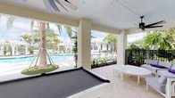 This single-level covered recreational pavilion offers a seamless blend of indoor comfort and outdoor living in a vibrant, resort-style setting. The space is anchored by a full-size billiards table positioned at the heart of the pavilion, complemented by a white round side table that adds a casual, social atmosphere. Overhead, a ceiling fan provides a cooling breeze, enhancing the open-air experience beneath the textured ceiling.

Large open-air columns frame sweeping, unobstructed views of the surrounding outdoor amenities, including a shimmering swimming pool lined with modern white lounge chairs. The pool area is beautifully landscaped with tall swaying palm trees and lush tropical greenery, creating a serene, vacation-like ambiance. A charming white pergola-style gazebo is visible in the background, adding to the resort aesthetic.

The flooring features classic light-toned tile, consistent with the bright and airy design of the space. Black metal fencing on one side adds a subtle boundary while preserving the open feel. Natural light floods the area throughout the day, making it an inviting and energetic gathering spot ideal for recreation and relaxation in a tropical community environment.