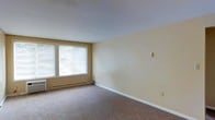 This single-floor residential space offers a clean, functional layout designed for comfortable everyday living. The interior is characterized by neutral beige walls that create a warm, understated backdrop throughout the space, complemented by wall-to-wall carpeting in a muted tone that adds softness underfoot and contributes to a quiet, cozy atmosphere.

Natural light plays a key role in the ambiance of the space. The living area features a wide double window fitted with horizontal blinds, allowing for adjustable light control while offering a view of the greenery outside. White trim and baseboards frame the room cleanly, providing a crisp contrast against the warm wall tones.

The space is equipped with baseboard heating units positioned along the exterior walls, ensuring consistent warmth throughout the colder months. The smooth, light-colored ceiling enhances the sense of openness within the room.

Overall, the space presents a straightforward and practical residential environment. Its neutral palette and carpeted floors make it a versatile canvas, easily adaptable to a variety of furnishing styles and personal preferences. The layout is simple yet efficient, offering a livable and well-lit interior suitable for a range of occupants.