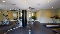 Located at 901 Kessler Ct in Garden City, Georgia, this single-floor fitness center is a well-equipped facility designed to support a comprehensive workout experience. The space is finished with durable dark flooring throughout, warm yellow-toned walls, and a textured ceiling fitted with recessed lighting and ceiling fans that maintain a comfortable environment during exercise.

The facility is generously outfitted with a variety of cardio and strength-training equipment, including elliptical machines, a treadmill, a stationary bike, a multi-station cable weight machine, a weight bench with free weights, and a rowing machine. Large wall-to-wall mirrors line one side of the room, visually expanding the space and allowing users to monitor their form during workouts.

Natural light flows in through multiple windows positioned along the perimeter walls, creating a bright and energizing atmosphere. A water cooler station is conveniently available for hydration, and informational charts and workout guides are posted on the walls. French-style glass doors provide access to the exterior, offering views of the surrounding greenery. A pair of seating chairs near the entrance adds a practical resting area. Overall, this fitness center is a thoughtfully arranged, fully functional space suited for a wide range of exercise routines.