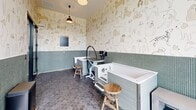 Located in Hyattsville, MD, this dedicated pet spa room is a thoughtfully designed grooming space that combines functionality with delightful, pet-friendly aesthetics. The room is wrapped in whimsical wallpaper featuring illustrated dogs of all breeds, creating a welcoming and playful atmosphere for both pets and their owners. A wainscoting of soft green glass mosaic tiles lines the lower half of the walls, complemented by hexagonal floor tiles that add texture and visual interest underfoot. The centerpiece of the space is a deep, built-in soaking tub with a step stool for easy pet access, alongside a hydraulic grooming table equipped with a professional arm and accessories. A built-in workstation with a dark stone countertop, undermount sink, and cabinetry provides a convenient prep and wash area, accented by a striking large-format wall print of a dog. A tall glass entry door floods the room with natural light and offers views of the surrounding greenery. Pendant lighting and wall-mounted sconces complete the warm, well-lit environment. Posted pet spa rules ensure a safe and orderly experience, reflecting the professional and caring nature of this uniquely appointed grooming space.
