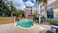 Located at 140 Holiday Blvd in Covington, Louisiana, this outdoor amenity space serves as a welcoming retreat set within a multi-story property. The centerpiece of the area is a generously sized freeform swimming pool with clear turquoise water, designed with gently curving edges that lend a resort-like aesthetic. A standout feature is the natural stone waterfall installation set against a wooden privacy wall, adding both visual interest and a soothing ambiance to the environment. The pool is framed by a spacious concrete sundeck equipped with lounge chairs and shaded umbrella seating, ideal for relaxation. Tall palm trees and lush tropical plantings are strategically placed throughout the space, reinforcing the Southern leisure atmosphere. The pool area is enclosed by black wrought iron fencing with sturdy column posts, providing both security and an open, airy feel. The surrounding multi-story building features a neutral stucco and stone facade with large windows that overlook the courtyard. A secondary seating area with additional lounge chairs and a market umbrella provides a quieter spot for guests. The overall design creates a comfortable, well-appointed outdoor space that reflects the warm, inviting character of southeastern Louisiana.