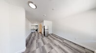 The Theadore Ada Studio in Tacoma, WA is a thoughtfully designed single-floor studio that maximizes every inch of its compact footprint. The open-plan layout seamlessly connects the living area and kitchen, creating a functional and cohesive living space. Durable patterned carpet tiles in neutral gray tones cover the main living and kitchen areas, lending a modern, low-maintenance aesthetic throughout.

The kitchen is equipped with stainless steel appliances, including a full-size refrigerator, microwave, and range, complemented by white cabinetry and an under-cabinet sink — providing all the essentials in an efficient configuration. A wall-mounted PTAC unit ensures year-round climate control, positioned conveniently beneath a window that allows natural light and outdoor views of the surrounding greenery.

The bathroom is designed with accessibility in mind, featuring sturdy grab bars near the toilet and shower area, a clean white color palette, and light wood-look flooring that adds warmth to the space. Flush-mount ceiling lights illuminate the unit throughout, maintaining a bright and welcoming ambiance. Clean white walls and simple, modern finishes create a neutral canvas throughout the studio, making it easy to personalize. The entry door provides direct access, adding to the unit's practical, straightforward layout.