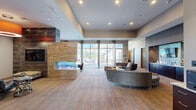 Located at 5265 Rockrose Ln in Allentown, PA, this thoughtfully designed community clubhouse offers a generous single-floor layout that seamlessly blends comfort, entertainment, and modern style. The entrance welcomes visitors with warm hardwood-style flooring, bold decorative plantings, and a large interior window overlooking a well-equipped fitness center, setting an inviting tone from the moment of arrival.

The main lounge is an expansive, light-filled gathering space anchored by a striking stone-clad double-sided fireplace. Curved sectional sofas and curated seating arrangements create an atmosphere ideal for both relaxed socializing and organized events. Floor-to-ceiling windows and glass doors bathe the room in natural light while providing direct views and access to an outdoor pool and patio area with umbrella-shaded seating.

Adjacent lounge nooks, adorned with framed vintage-style artwork, offer more intimate settings with leather recliner chairs and bistro tables — perfect for casual conversation or quiet relaxation.

The entertainment room is a standout feature, centering on a large granite-topped bar island with bar stool seating, pendant lighting, and a sleek wet bar with mosaic tile backsplash and mounted television. A pool table and additional high-top seating round out this dynamic recreation space. Throughout, the design maintains a cohesive contemporary aesthetic with warm neutral tones, rich dark wood accents, and carefully layered lighting.