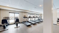 Located at 14504 Abbeyfield Ave in Rosemount, MN, this fitness center offers a well-appointed, single-floor exercise space designed to support a comprehensive workout routine. The exercise room is generously sized and divided into distinct functional zones, each catering to different training needs.

The cardio section is well-stocked with a row of Spirit treadmills, stationary bikes, and elliptical machines, all positioned along a wall of large windows that invite abundant natural light and provide open outdoor views — an energizing backdrop for any workout. The flooring in this area consists of durable, textured rubber matting, well-suited for high-traffic use.

A separate strength training area features a multi-station cable and weight machine, offering a variety of resistance exercises. An additional open zone with smooth hardwood-style flooring provides space for a rowing machine and flexible floor-based activities such as stretching or bodyweight training.

The facility also includes a water fountain station, ensuring convenience during workouts. Throughout the space, a drop ceiling with recessed lighting maintains a clean, modern aesthetic. Neutral wall tones in warm beige and white contribute to a bright, motivating atmosphere. The layout is thoughtfully organized, making efficient use of the available space while maintaining clear circulation throughout.