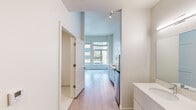 Located at 1741 36th St in Denver, Colorado, this single-floor unit presents a clean, contemporary design characterized by crisp white walls, light wood-tone flooring, and an open, airy layout. The space benefits from notably tall ceilings throughout, enhancing the sense of openness, with an exposed metal duct element adding an industrial touch to the main living area.

The kitchen is efficiently designed and well-appointed, featuring stainless steel appliances including a refrigerator, electric range, and over-the-range microwave. White cabinetry pairs with light countertops, and open shelving above the sink provides additional storage. A peninsula counter extends the workspace and offers a casual dining or prep area.

Large windows in the main living area flood the space with natural light and provide views of the surrounding Denver streetscape. The bathroom is finished with neutral tile flooring, a tiled walk-in shower, and chrome fixtures, maintaining the unit's consistent clean aesthetic. A separate door leads to a tiled entryway area. Track lighting throughout ensures consistent, adjustable illumination. The overall ambiance is modern, minimal, and functional.