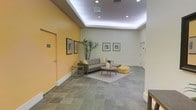 This single-floor space presents a well-designed lobby or communal lounge area that combines functional design with inviting aesthetics. The room is immediately distinguished by its bold yellow accent wall, which contrasts warmly against the surrounding sage-green and neutral tones, creating a vibrant yet balanced atmosphere. The ceiling features a recessed tray design outlined with ambient LED lighting, adding depth and a polished architectural detail to the space.

The flooring consists of large-format stone-look tiles in muted gray tones, lending a durable and sophisticated foundation throughout. A central seating arrangement anchors the lounge area, featuring a contemporary gray sofa, an ottoman in a matching yellow hue, and a round wooden coffee table atop a patterned area rug, tying the color palette together cohesively.

Framed artwork lines the walls, adding a curated, gallery-like quality to the space. A tall indoor plant introduces a natural element, softening the clean architectural lines. Recessed ceiling lighting provides even illumination throughout, complementing the ambient tray lighting. An access door with a room number plate is visible along the yellow wall, suggesting this space serves as a shared common or reception area.