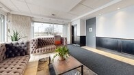 Located at 333 Rue Riverside in Saint-Lambert, Quebec, this well-appointed building lobby offers a welcoming and polished first impression across a single floor. The space is divided into two distinct areas, each thoughtfully designed to blend functionality with comfort.

The first area features rich, dark wood wainscoting paired with textured patterned wallcovering, recessed accent lighting, and a cozy seating arrangement of upholstered orange armchairs centered around a side table. A wall-mounted flat-screen television and a decorative mirror with greenery add character, while elevator access is conveniently positioned nearby.

The second area transitions to a more contemporary aesthetic, anchored by floor-to-ceiling windows that flood the space with natural light and offer open views of the surrounding streetscape. A tufted Chesterfield-style sofa, a metal-framed coffee table, and an ornate area rug create an inviting lounge atmosphere. A textured stone accent wall, a modern built-in display unit, and vibrant potted plants contribute to the space's refined yet approachable ambiance.

The entry vestibule features glass doors, bold red planters, a mailbox wall, and additional greenery, establishing a clean and organized arrival experience. Tile and carpet flooring zones delineate pathways throughout, while a drop ceiling with recessed lighting maintains a consistent, polished finish across the entire lobby.