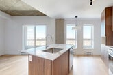 Located at 4845 Chemin de la Côte-Saint-Luc in Montréal, this thoughtfully designed one-bedroom, one-bathroom unit offers a refined contemporary living experience. The open-concept layout seamlessly connects the kitchen and living area, bathed in natural light from large windows and a sliding glass door that frames expansive outdoor views. Light hardwood flooring runs throughout, complementing the clean white walls and exposed concrete ceiling and structural columns — a striking architectural contrast that lends the space an urban, loft-inspired character. The well-appointed kitchen serves as the heart of the unit, anchored by a central island with a white quartz countertop, an undermount stainless steel sink, and warm wood cabinetry paired with greige upper cabinets. Stainless steel appliances, including a refrigerator and range, complete the functional setup, while a decorative backsplash adds a refined finishing touch. The bedroom is a calm, well-proportioned retreat featuring the same hardwood flooring, exposed concrete ceiling, a bright window with open views, and a double sliding-door closet. The bathroom offers a clean, modern design with wood-toned vanity cabinetry, a mirrored medicine cabinet, and a glass-enclosed shower area with crisp white finishes throughout.