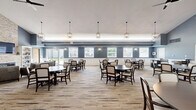 Located in Shelby Township, Michigan, this generously sized single-floor community space offers a welcoming and well-appointed environment designed for comfort and social interaction. The interior is finished with a consistent palette of cool slate-blue walls, crisp white wainscoting, and durable wood-look plank flooring that flows seamlessly throughout.

The living room serves as a focal point, anchored by a floor-to-ceiling stacked stone fireplace with a mounted television and flanked by built-in white bookshelves. Vaulted ceilings amplify the sense of openness, while plush seating and an industrial-style chandelier contribute to a warm, inviting ambiance. French doors provide direct access to the outdoors, allowing natural light to pour into the space.

The kitchen is outfitted with rich dark-wood cabinetry, granite countertops, a mosaic tile backsplash, and stainless steel appliances, including a microwave and dishwasher. A deep sink and ample counter space make it well-suited for group gatherings and events.

The dining area accommodates multiple round tables with upholstered seating, ideal for communal dining. Large windows line the walls, offering views of the surrounding community and filling the room with natural light. Wall sconces and pendant lighting throughout add a refined, polished finish to the overall design.
