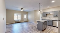 Located at 2067 11th Ave E in Menomonie, Wisconsin, this thoughtfully designed single-story home offers three bedrooms and three full bathrooms within a well-utilized floor plan. The open-concept kitchen and living area serves as the heart of the home, highlighted by gray shaker-style cabinetry, granite-look countertops, a central island with pendant lighting, and a full suite of stainless steel appliances including a refrigerator, range, microwave, and dishwasher. Light wood-look luxury vinyl plank flooring runs throughout the main living areas, complementing the neutral warm-beige walls and crisp white trim. The living room benefits from natural light through large windows overlooking the surrounding landscape, and a ceiling fan adds comfort to the space. Bedrooms feature plush carpeting, ceiling fans, and windows with outdoor views, while the generously sized primary bedroom includes a walk-in closet with wire shelving. Each bathroom is finished with gray vanity cabinetry, matte black hardware, light countertops, and matching LVP flooring. A dedicated utility room provides additional functional space. The home's clean, contemporary aesthetic and efficient single-floor layout make it a practical and comfortable residence.