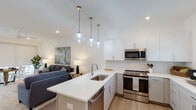 Located at 4550 Atrium Ct in Wilmington, NC, this contemporary two-story home offers a thoughtfully designed layout with clean lines and modern finishes throughout. The open-concept main level centers around a bright living area with vaulted ceilings, recessed lighting, and a ceiling fan, creating an airy and welcoming atmosphere. The kitchen is a standout feature, boasting crisp white upper cabinetry, two-tone lower cabinetry, stainless steel appliances including a refrigerator, range, and over-the-range microwave, a large white quartz island with an undermount sink, and globe pendant lighting. Light wood-style flooring flows seamlessly throughout the main level. A staircase leads to the upper floor, with a convenient bathroom and additional living space visible on the lower level. The home includes multiple well-appointed bedrooms, each with ceiling fans and natural light from large windows. The primary bathroom features a long white vanity, a frameless glass walk-in shower, and elegant lighting, while a secondary bathroom offers a full tub-shower combination. Each room reflects a cohesive, modern coastal aesthetic suited for comfortable everyday living.