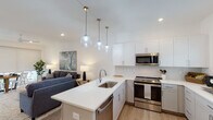 Located at 4550 Atrium Ct in Wilmington, NC, this contemporary two-story home offers a thoughtfully designed layout with clean lines and modern finishes throughout. The open-concept main level centers around a bright living area with vaulted ceilings, recessed lighting, and a ceiling fan, creating an airy and welcoming atmosphere. The kitchen is a standout feature, boasting crisp white upper cabinetry, two-tone lower cabinetry, stainless steel appliances including a refrigerator, range, and over-the-range microwave, a large white quartz island with an undermount sink, and globe pendant lighting. Light wood-style flooring flows seamlessly throughout the main level. A staircase leads to the upper floor, with a convenient bathroom and additional living space visible on the lower level. The home includes multiple well-appointed bedrooms, each with ceiling fans and natural light from large windows. The primary bathroom features a long white vanity, a frameless glass walk-in shower, and elegant lighting, while a secondary bathroom offers a full tub-shower combination. Each room reflects a cohesive, modern coastal aesthetic suited for comfortable everyday living.