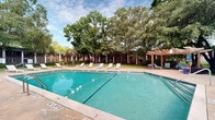 Located at 5600 Dorchester Rd in North Charleston, South Carolina, this outdoor amenity area centers around a generously sized rectangular swimming pool with clearly marked shallow and deep ends ranging from 3 to 7 feet. The pool features clean, turquoise-tinted water, lane dividers, and easy-access stainless steel entry rails. Surrounding the pool is a spacious concrete deck furnished with multiple white lounge chairs, providing ample space for sunbathing and relaxation. A wooden pergola structure situated at one end of the deck offers a shaded gathering spot, adding architectural character to the outdoor environment. A freestanding umbrella table provides additional shade on the opposite side of the pool area. The space is enveloped by a canopy of large, mature trees — including oaks and palms — that create a lush, park-like atmosphere and offer natural shade throughout the day. Well-maintained green lawn surrounds the perimeter, enhancing the overall sense of openness. Brick residential buildings are visible in the background, integrating the pool area seamlessly within the broader community. The combination of generous pool dimensions, thoughtful seating arrangements, and natural landscaping makes this an inviting and functional outdoor recreational space.