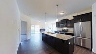 This single-floor residence is part of a modern brick building and showcases a thoughtfully designed open-concept layout. The kitchen serves as a central highlight, featuring dark cabinetry that contrasts elegantly with light granite countertops. A large central island with an undermount sink and pendant lighting overhead provides both functional workspace and a natural gathering point. Stainless steel appliances, including a range and built-in microwave, complete the well-appointed kitchen.

The kitchen flows seamlessly into the living area, which benefits from abundant natural light streaming through large sliding glass doors and windows. A sleek, modern linear fireplace anchors the living space, adding warmth and visual interest. Dark hardwood flooring extends throughout, tying the open-concept space together cohesively.

The exterior of the building features classic red brick construction with structured balconies and white railings. A generously sized shared outdoor terrace is finished with lush artificial turf and a paved pathway, offering a comfortable outdoor area with open views of the surrounding landscape and tree line beyond the railing perimeter.