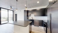 This well-appointed one-bedroom, one-bathroom unit showcases a contemporary industrial aesthetic throughout. Exposed concrete ceilings and structural columns contrast elegantly with crisp white walls and light-toned hardwood flooring, creating a sophisticated urban atmosphere. The open-concept kitchen and living area serves as the heart of the unit, featuring dark flat-front cabinetry, a white subway tile backsplash, stainless steel appliances including a French door refrigerator, gas range, built-in microwave, and dishwasher. A large central island with a charcoal quartz countertop provides additional prep space and casual seating, illuminated by stylish globe pendant lights. The living area opens to a private balcony through full-height sliding glass doors, offering expansive city views and abundant natural light. The bedroom continues the industrial-modern theme with exposed concrete accents, a ceiling fan, and a floor-to-ceiling window framing urban vistas. A dedicated closet provides organized storage. The bathroom is finished with dark cabinetry, a gray quartz vanity, a framed mirror, matte black hardware, and a tiled walk-in shower. Throughout, bold black interior doors add a cohesive design detail that ties the space together.