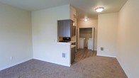 This single-floor unit features an open-concept layout that seamlessly connects the living area to the kitchen. The living space is finished with wall-to-wall carpeting, contributing to a warm and comfortable atmosphere, while the kitchen area transitions to hard-surface flooring for easy maintenance. A half-wall partition defines the kitchen zone without fully closing it off from the main living area, preserving an open and airy feel throughout.

The kitchen is equipped with dark-toned cabinetry that provides a subtle contrast against the light-colored walls, along with a built-in cooktop. Flush-mount ceiling lighting illuminates both the kitchen and the adjacent hallway area effectively.

Toward the rear of the unit, a dedicated in-unit laundry area houses side-by-side washer and dryer units, adding significant convenience. Adjacent to the laundry space is a walk-in closet, offering generous storage capacity. The unit also features floor-level HVAC vents integrated into the walls, ensuring efficient climate control throughout. The overall palette of neutral tones and clean lines gives the space a simple, functional character.