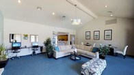 This single-story space is designed with an open-concept layout that maximizes its sense of spaciousness and flow. The centerpiece of the main living area is a dramatic vaulted ceiling with exposed beams, creating a bright and airy atmosphere. A modern pendant light fixture hangs as a focal point above the seating arrangement, which features a large sectional sofa and coordinating ottoman in neutral tones, accented with teal and patterned throw pillows that add a coastal-inspired character.

Soft blue carpeting extends throughout the main living area, grounding the space with a calming, cohesive feel. A dedicated workstation in the corner is equipped with a desktop computer, making the space functional alongside its comfortable design. Lush potted plants are thoughtfully placed throughout, bringing life and texture to the interior.

Framed artwork adorns the walls near a dining area, which is visible in the background alongside a compact, well-appointed kitchen featuring warm wood cabinetry and bar-height seating. Natural light filters through multiple windows, enhancing the open and welcoming character of the space. Overall, this versatile, single-floor layout blends comfort, practicality, and style seamlessly.