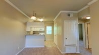 This single-floor residence offers a welcoming and functional layout highlighted by an open-concept design that seamlessly connects the kitchen and living area. The kitchen is outfitted with white cabinetry, white appliances, and a peninsula-style breakfast bar with a countertop overhang, making it ideal for casual dining and everyday use. A stylish pendant light hangs above the kitchen area, adding a decorative focal point to the space. Crown molding runs along the ceiling, lending a touch of classic architectural detail throughout. The living area features plush carpeting in warm neutral tones, creating a cozy and comfortable atmosphere. Tile flooring in the kitchen and adjacent areas provides a practical, easy-to-clean transition. A window positioned near the kitchen allows natural light to filter in, brightening the interior and offering a glimpse of lush greenery outside. A full-panel white door near the entryway adds clean, classic character to the space. The overall color palette of soft beige and white tones creates a cohesive and inviting environment, well-suited for comfortable everyday living. The single-floor layout ensures ease of navigation throughout the home.