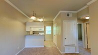This single-floor residence offers a welcoming and functional layout highlighted by an open-concept design that seamlessly connects the kitchen and living area. The kitchen is outfitted with white cabinetry, white appliances, and a peninsula-style breakfast bar with a countertop overhang, making it ideal for casual dining and everyday use. A stylish pendant light hangs above the kitchen area, adding a decorative focal point to the space. Crown molding runs along the ceiling, lending a touch of classic architectural detail throughout. The living area features plush carpeting in warm neutral tones, creating a cozy and comfortable atmosphere. Tile flooring in the kitchen and adjacent areas provides a practical, easy-to-clean transition. A window positioned near the kitchen allows natural light to filter in, brightening the interior and offering a glimpse of lush greenery outside. A full-panel white door near the entryway adds clean, classic character to the space. The overall color palette of soft beige and white tones creates a cohesive and inviting environment, well-suited for comfortable everyday living. The single-floor layout ensures ease of navigation throughout the home.