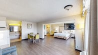 Located at 1400 W Tunnel Blvd in Houma, Louisiana, this well-appointed hotel suite offers a comfortable and functional layout across a single floor. The expansive bedroom is the centerpiece of the suite, featuring wood-style flooring, a textured ceiling, large windows with drape treatments that allow ample natural light, a wall-mounted flat-screen TV, and individual climate control units. A seating area with a sofa complements the sleeping space, adding to the room's versatility.

The suite includes four bathrooms, each finished with warm beige tile work, tub-shower combinations with curtains, and clean, well-lit vanity areas. The vanities feature wide countertops with integrated sinks, framed mirrors, bright overhead lighting, and practical amenities such as hair dryers, towel hooks, and luggage racks — all reflecting a thoughtful hospitality design.

A dedicated dining area just off the main living space features a rectangular table with four upholstered chairs in a chartreuse tone, adding a pop of color against the neutral walls. Subtle decorative wall art enhances the welcoming atmosphere. The overall design is clean and functional, catering to guests seeking a comfortable extended-stay or short-term lodging experience in Houma, Louisiana.