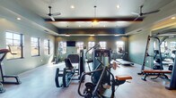 Located at Forest Village Drive in Holly Springs, NC, this fitness center is a thoughtfully designed, single-story facility offering a comprehensive workout environment across several distinct zones. The main strength training area is anchored by an array of cable machines, weight equipment, and resistance stations, all set against deep sage-green walls that lend the space a modern, upscale aesthetic. Exposed wooden ceiling beams, recessed lighting, and industrial-style pendant lights contribute to a warm yet energetic atmosphere throughout.

A dedicated cardio zone accommodates multiple treadmills, elliptical trainers, and an assault bike, positioned alongside large windows that allow generous natural light and outdoor views of the surrounding landscape. An additional smaller room provides space for a spin bike and other individual training equipment, benefiting from abundant window coverage on multiple walls.

The facility also includes a separate studio room finished in teal-accented walls, outfitted with a ballet barre, floor-to-ceiling mirrors, a wall-mounted TV, and open floor space suited for group fitness, yoga, or stretching routines. Glass-paneled doors with transom windows connect the studio to the main fitness floor, maintaining visual openness. Ceiling fans throughout ensure comfortable airflow, and light-toned flooring unifies the overall design.