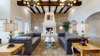 This elegantly designed single-floor community space blends classic Southern charm with refined interior details throughout. The grand lounge serves as the focal point, boasting soaring vaulted ceilings with rich dark exposed wood beams, dramatic wrought-iron chandeliers, and a full-height exposed brick accent wall anchored by a stately white-painted fireplace. Tufted leather Chesterfield sofas flank an ornate iron-and-marble coffee table, all set atop richly patterned encaustic tile flooring that extends into the adjacent dining area. The dining room features a coffered ceiling with large pendant drum lights, wainscoting, framed botanical art, and multiple round and rectangular tables with cross-back chairs upholstered in soft blue. Dark-framed French doors with glass panes connect the various areas, creating a cohesive yet defined flow throughout the space. The full kitchen is outfitted with white shaker cabinetry, quartz countertops, a bold black-and-white patterned tile backsplash, stainless steel appliances including a French door refrigerator and double wall ovens, and a substantial center island. A secondary service counter area features white cabinetry, an arched mirror, and a utility sink. Wide-plank hardwood flooring, abundant natural light, and automatic glass entry doors completing the welcoming, community-oriented atmosphere.