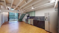 This two-story loft in Rogers, Arkansas, blends industrial character with modern updates across two well-defined levels. The main floor opens with a striking open-plan layout showcasing warm hardwood floors and soaring exposed beam ceilings painted white, lending a clean, airy industrial aesthetic. A large grid-pane window wall floods the space with natural light and offers views to the exterior. The kitchen anchors the main floor with sage-green cabinetry, a butcher block countertop, gray subway tile backsplash, and a full suite of stainless steel appliances. A statement staircase with green-painted treads and cable-rail detailing connects the two levels, serving as both a functional and architectural focal point. Exposed brick accents add raw texture and warmth to the main living area. The upper level provides a comfortable open room with light hardwood flooring, a white-painted exposed beam ceiling, a wall-mounted mini-split HVAC unit, and a ceiling fan. A well-appointed walk-in closet with dual hanging rods and shelving offers generous storage. The bathroom features floor-to-ceiling white subway tile, a glass-enclosed walk-in shower with matte black fixtures, a vessel sink on a wood-and-metal vanity table, and penny tile flooring — a refined, design-forward finish throughout.
