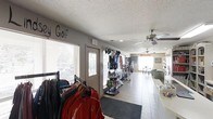 This golf pro shop provides a functional and well-organized retail environment for golf enthusiasts. The single-floor layout is designed to maximize product visibility and ease of navigation for customers. Upon entry, clothing racks prominently display a variety of golf apparel, including jackets and shirts from recognizable brands, making it easy to browse a curated selection of on-course attire. Centrally positioned display racks showcase an assortment of golf accessories and equipment, keeping merchandise accessible throughout the space. The flooring is composed of durable tile, transitioning to a mat near the entrance for practicality. Ceiling fans are installed throughout, contributing to comfortable air circulation across the retail floor. Recessed lighting complements the natural light brought in by windows along the front of the shop, creating a bright and inviting atmosphere. Toward the rear of the space, a counter area provides a dedicated point of sale and customer service zone, while a seating area offers a comfortable spot for visitors. Wall-mounted shelving near the back provides additional display capacity for merchandise and informational materials. The textured ceiling and neutral wall tones give the shop a clean, approachable aesthetic suited to its purpose as a hub for golfers seeking quality gear and attire.